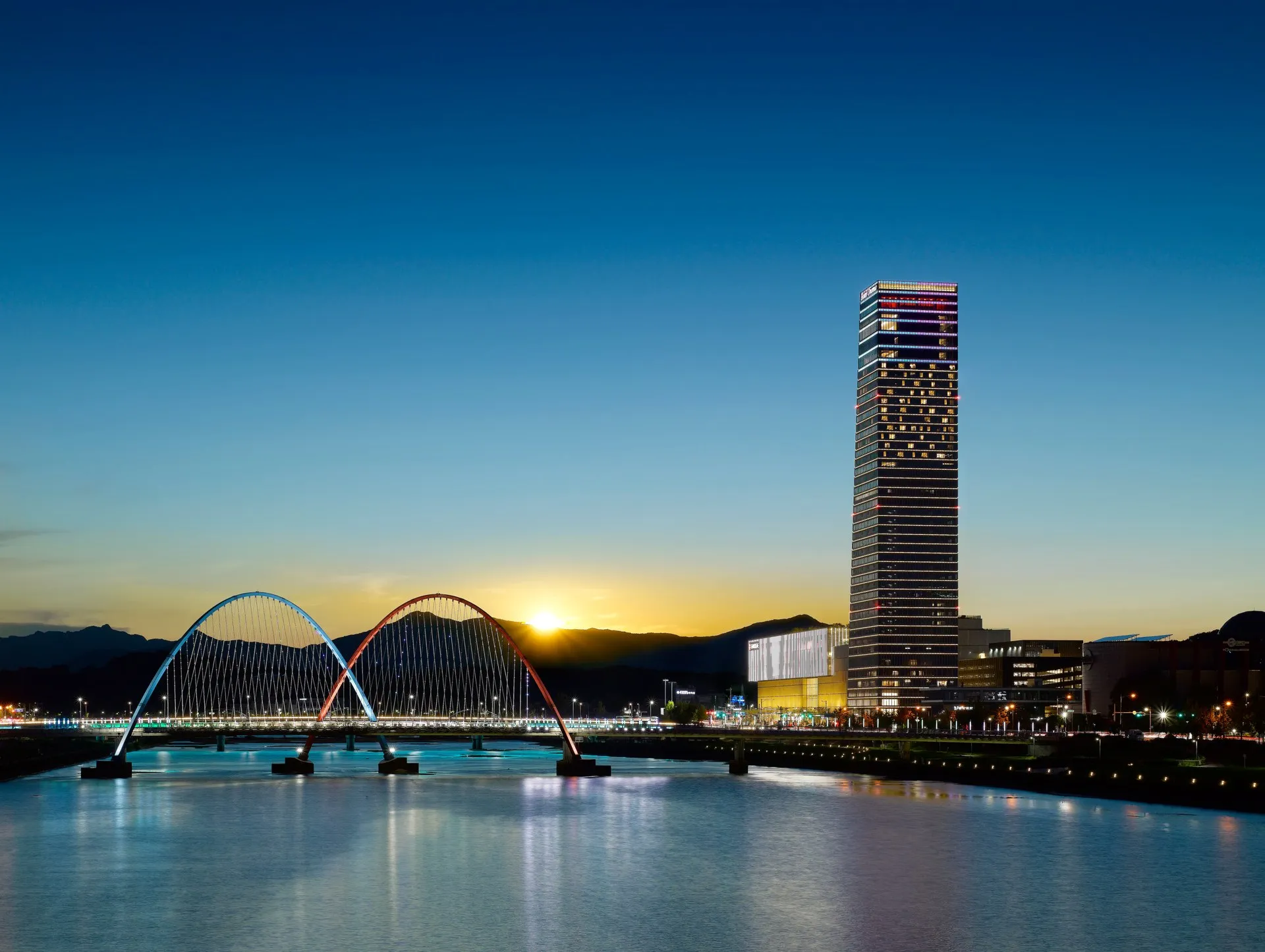 The Expo Bridge in Daejeon at dusk, its lit arches reflected in the river below the city skyline.